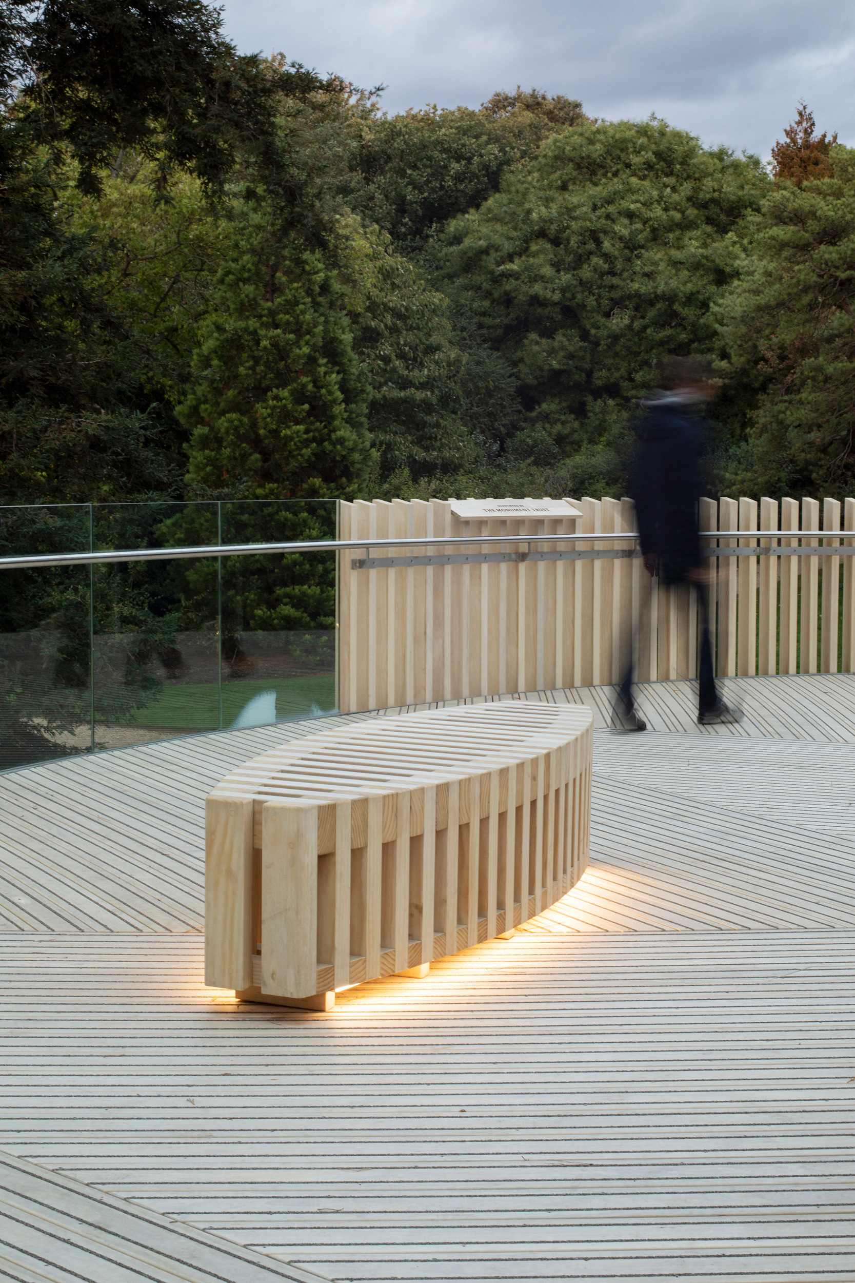 Under seat lighting used on a bench on the viewing platform of The Rising Path Cambridge University Botanic Garden by architects CDC Studio. Blurred shape of a Visitor in the background.
